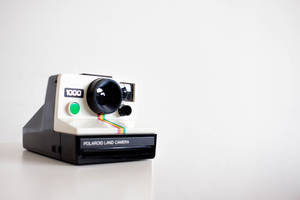 一台復古Polaroid Land Camera 1000相機，帶有彩虹底片條，置於白色表面上。 — 拍立得 壁紙