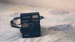 花卉圖案床罩上的黑色Polaroid Sun 660相機。 — 拍立得 壁紙