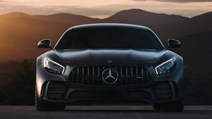 啞光黑色Mercedes-AMG GT在夕陽下對著鏡頭，背景有山脈。 — Mercedes-Benz 壁紙