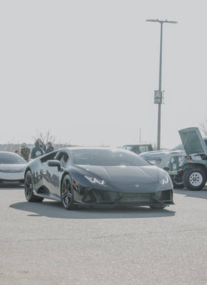 消光黑色藍寶堅尼 Huracán 停放在陽光明媚的停車場，附近有人群。 — 蘭博基尼 壁紙