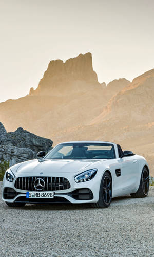 白色 Mercedes-AMG GT Roadster 停泊在碎石上，靠近岩石山脈，日落時分。 — Mercedes Amg iPhone 壁紙