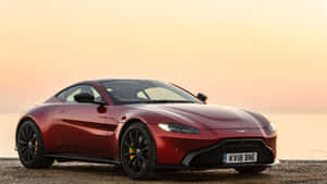 紅色Aston Martin跑車停放在日落時分的鵝卵石海灘上。 — 奧斯頓馬丁 壁紙