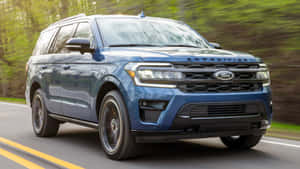 藍色福特 Expedition SUV 在穿過林區的柏油路上行駛。 — 福特 壁紙