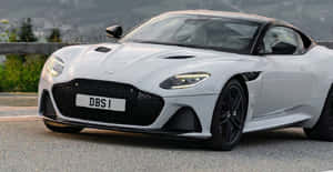 銀色Aston Martin DBS1跑車在山路上，背景有翠綠山丘。 — 奧斯頓馬丁 壁紙
