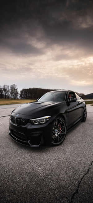 黑色BMW M2雙門跑車停在裂開的柏油路上，背景是暴風雨黃昏的天空。 — iPhone 17e 壁紙