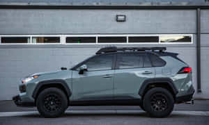 鼠尾草綠色豐田RAV4，搭配黑色配件，停在灰色磚造建築前。 — 低調 壁紙