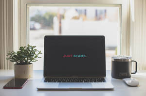 螢幕顯示「JUST START.」的筆記型電腦，旁邊有植物、杯子和電話在窗台上。 — 激勵 壁紙