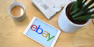 平板電腦顯示eBay標誌在木桌上，附有咖啡杯、報紙和植物。 — eBay 壁紙