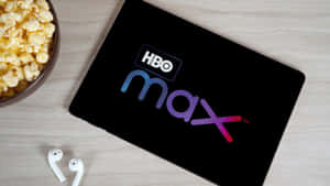 平板電腦帶有HBO Max標誌，旁邊有爆米花和耳塞，在木桌上。 — 低調 壁紙