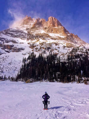 一名滑雪者站在雪地上，面對著一座雄偉的山峰，下方有常青樹。 — 冬季風景 壁紙