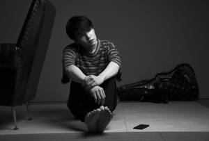 年輕男子赤腳坐在地板上，旁邊有吉他盒和椅子。