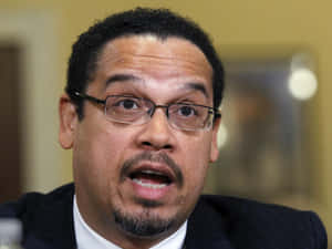 穿西裝戴眼鏡的男子在正式會議中發言。 — 政治 壁紙