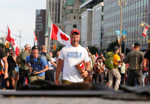 戴紅色帽子和身穿Canada Marches襯衫的男子，在日間抗議活動中手持裱框照片，背景有加拿大國旗和城市建築。 — 慶祝 壁紙