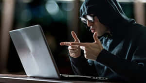 一名穿著黑色連帽衫和護目鏡的人在黑暗的房間中指向筆記型電腦螢幕。 — 駭客面具 壁紙