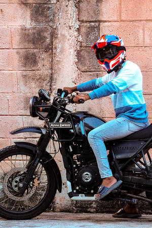 在磚牆附近，一名戴著彩色頭盔的人騎在 Royal Enfield 摩托車上。