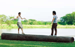 兩人在湖邊的木頭上保持平衡，背景有綠樹。 — 花園 壁紙