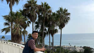 男子在陽臺上微笑，身後是海洋和棕櫚樹 — 海灘度假 壁紙