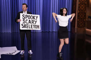 男子舉著spooky scary skeleton牌子，女子在舞台上跳舞。 — 大笑 壁紙
