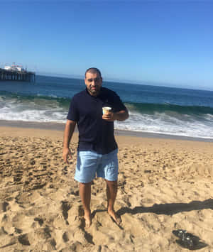 陽光明媚的海灘上，男子在碼頭附近手持咖啡，伴隨著海洋波浪。 — 海灘 壁紙