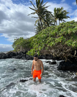 穿著橙色泳褲的男子在熔岩岩石和棕櫚樹附近的海浪中涉水。 — 海洋 壁紙