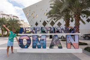 男子在棕櫚樹下與Experience Oman標誌合影 — 棕櫚樹 壁紙