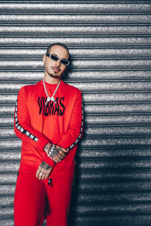 身穿紅色運動服和太陽眼鏡的男子靠在金屬百葉窗上。 — J Balvin 壁紙