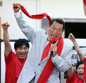 身穿灰色西裝、打紅色領帶的男子對著麥克風大喊，舉起拳頭和紅絲帶。 — 運動 壁紙