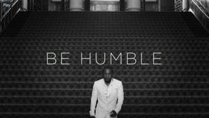 身穿白色西裝的男子站在華麗樓梯底部，帶有「BE HUMBLE」文字。