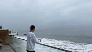 身穿白色運動衫的男子在多雲的日子裡從現代玻璃陽台凝視海洋。 — 海灘 壁紙