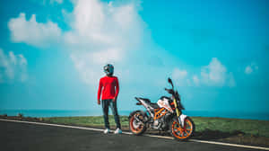 身穿紅色襯衫的摩托車手站在沿海公路上，旁邊是一輛橙色和白色的摩托車。 — 藍天 壁紙