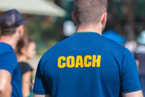 男士身穿藍色襯衫，印有黃色COACH文字，在戶外陽光下。 — 教練 壁紙