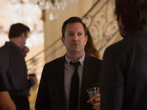 身穿西裝的男子在設有華麗樓梯和吊燈的正式室內派對中手持飲品。 — 約翰·藍儂 GIF 壁紙