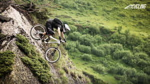 山地車手在陡峭懸崖小徑上騰空，俯瞰綠色山谷。 — 登山車 壁紙