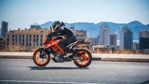 機車騎士騎橙色KTM Duke在城巿道路上，背景有山巒 — 巴布風機車 壁紙