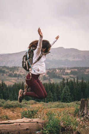 一名背著背包的女子在樹木和山丘環繞的山間草地上跳躍。 — 秋季 壁紙