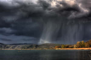 黑暗的暴風雲將雨水傾瀉在一個平靜的湖面上，遠處有山丘和樹木。 — 閃電麥坤 壁紙