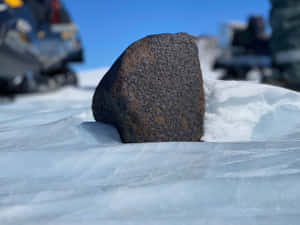 一塊深色岩石從藍冰中突出，背景有雪地摩托車。 — 南極洲 壁紙