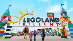 家庭在LEGO Billund主題公園入口微笑，周圍有色彩繽紛的LEGO結構。 — 樂高 壁紙
