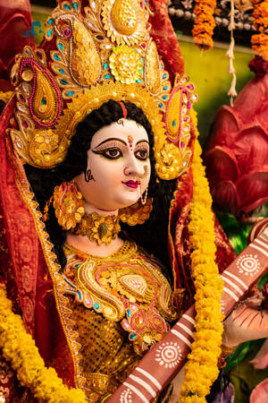 一尊裝飾著金色珠寶和萬壽菊花環的鮮豔印度教女神雕像。 — Maa Durga 壁紙