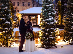 一對情侶在夜晚的雪地公園中，靠近點亮的樹木站立。 — 可愛聖誕快樂 壁紙