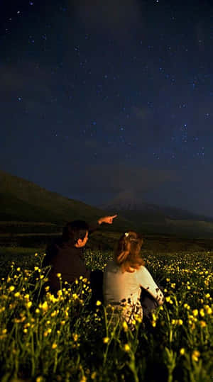 兩個人坐在黃色花田裡，指著山上的星星。 — 夜空 壁紙