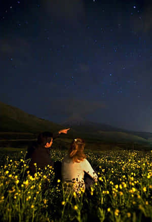 兩個人坐在黃色花田裡，指著山上的星星。 — 夜晚 壁紙