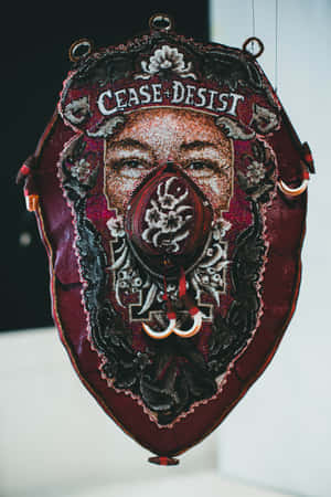 掛在牆上的刺繡紡織藝術品，展示了一張帶有「CEASE DESIST」文字的臉孔。 — 盾之勇者 壁紙