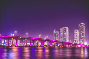 夜晚中，一座發光的紫色橋樑橫跨水面，城市燈光倒映其中。
