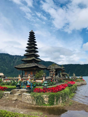 湖邊的峇里島寺廟，周圍環繞著紅花和綠色山丘，天空多雲 — 廟宇 壁紙