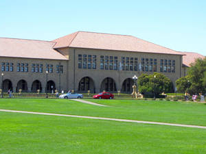 一座大型大學建築，帶有拱形窗戶和赤陶屋頂，位於綠色草坪後面，草坪上停有汽車和行人。 — 草地 壁紙