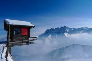 一座紅色門的木造山間小屋，位於雲端與積雪山峰之上。 — 寒冷 壁紙