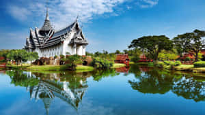 白色泰式寺廟倒映在平靜的池塘中，周圍有綠樹和藍天 — 海灘桌面 壁紙