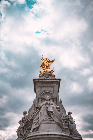 一座金色天使雕像在陰雲密布的天空下矗立於石碑之上。 — 伊莉莎白女王 壁紙