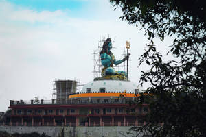 被樹枝環繞的寺廟圓頂上，一座正在建造中的大型藍色濕婆神像。 — 斯瓦米納拉揚 壁紙